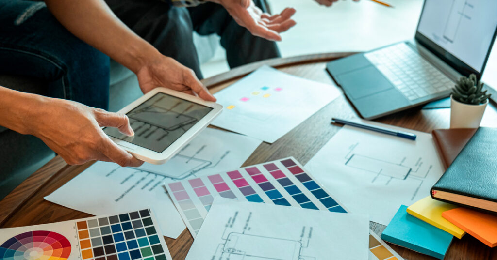 Dos personas hablando sobre como hacer branding efectivo y una mesa llena de elementos relacionados con el branding, como paletas de colores, un macbook y un ipad, post-its, agendas, etc